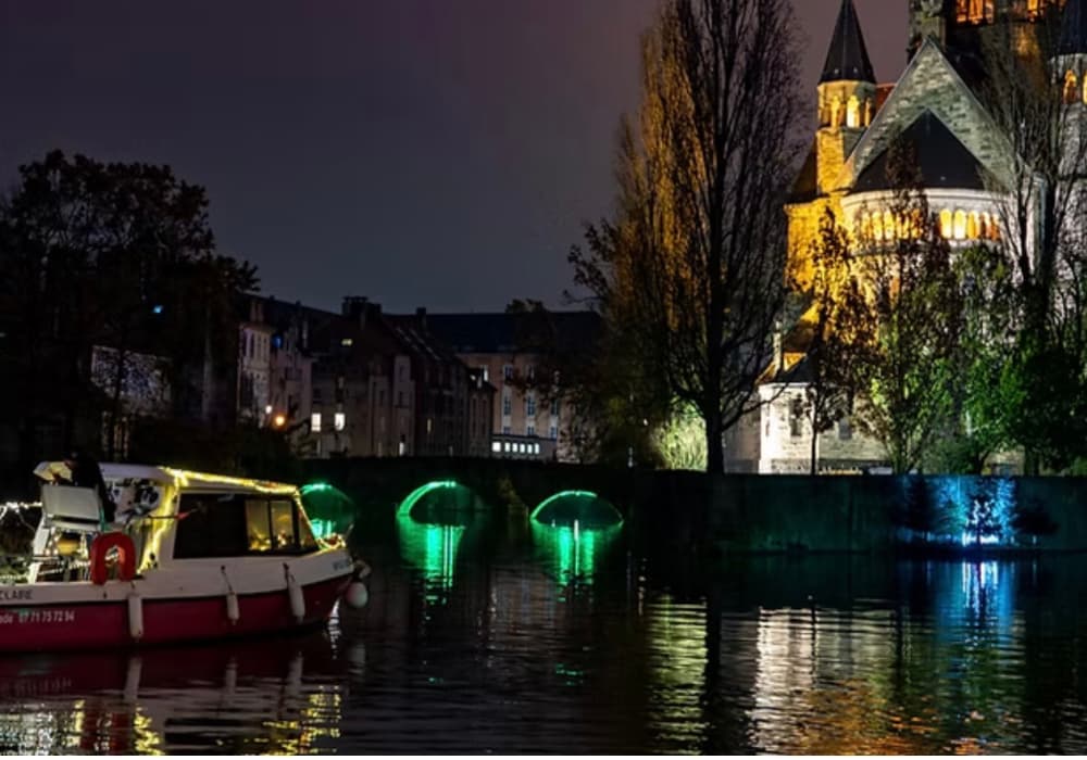 bateau illuminé naviguant à Metz avec reflets sur l’eau et vue sur le temple Neuf