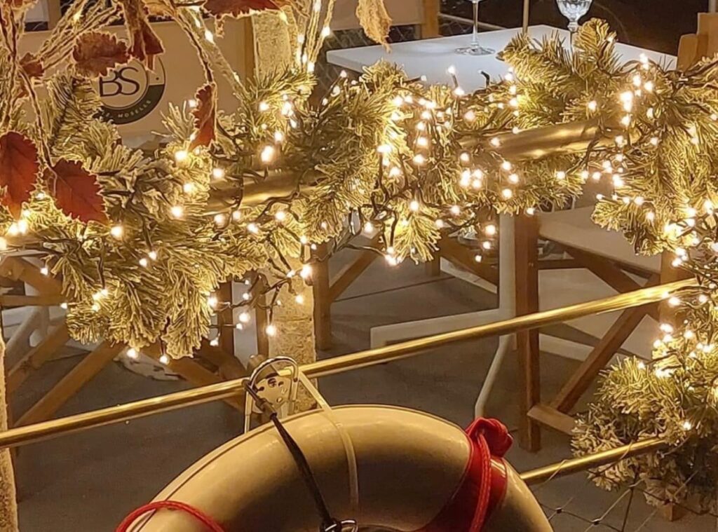 Main courante d'un bateau décorée d'une épaisse guirlande de sapin illuminée et bouée de sauvetage au premier plan.